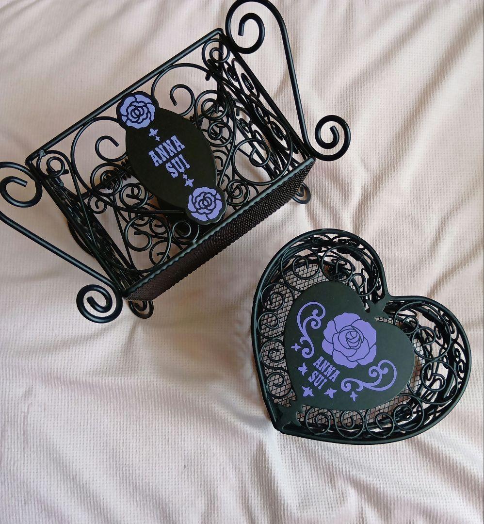 Two black decorative steel small storage boxes with ANNA SUI logo and purple rose motifs shown on a light fabric surface.