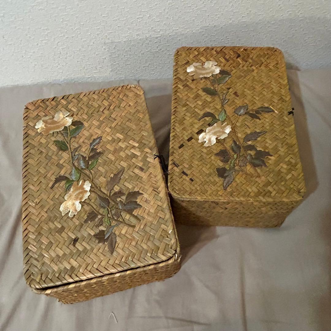 Two antique straw baskets with floral embroidery placed side by side on a fabric surface.