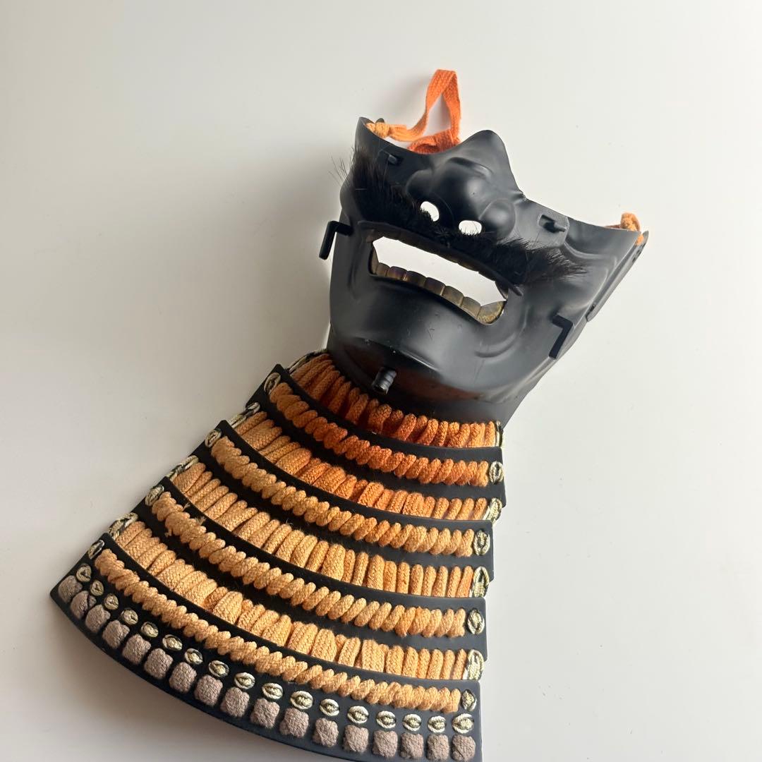 Wooden Japanese Noh menpo face mask with orange cords and an orange-and-black segmented throat guard laid on a white background.