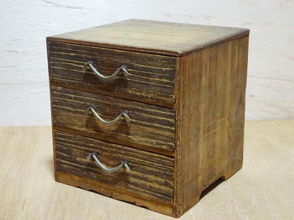 Wooden three-drawer small chest with metal handles shown at an angle on a wooden surface.