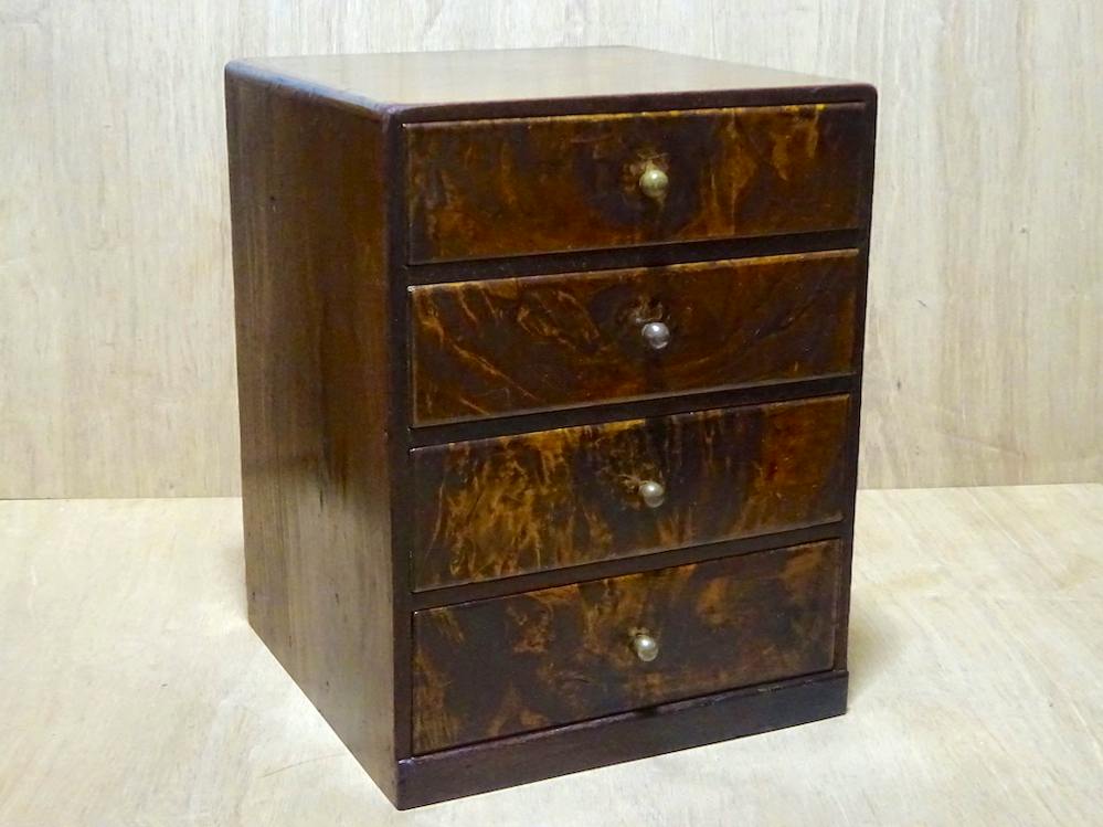 Small four-drawer wooden chest with dark patterned drawer fronts photographed on a light wood surface.