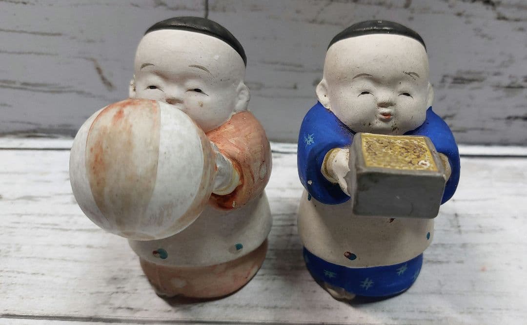 Front view of two ceramic child figurines on a wooden surface, the left holding a ball and the right holding a small box.