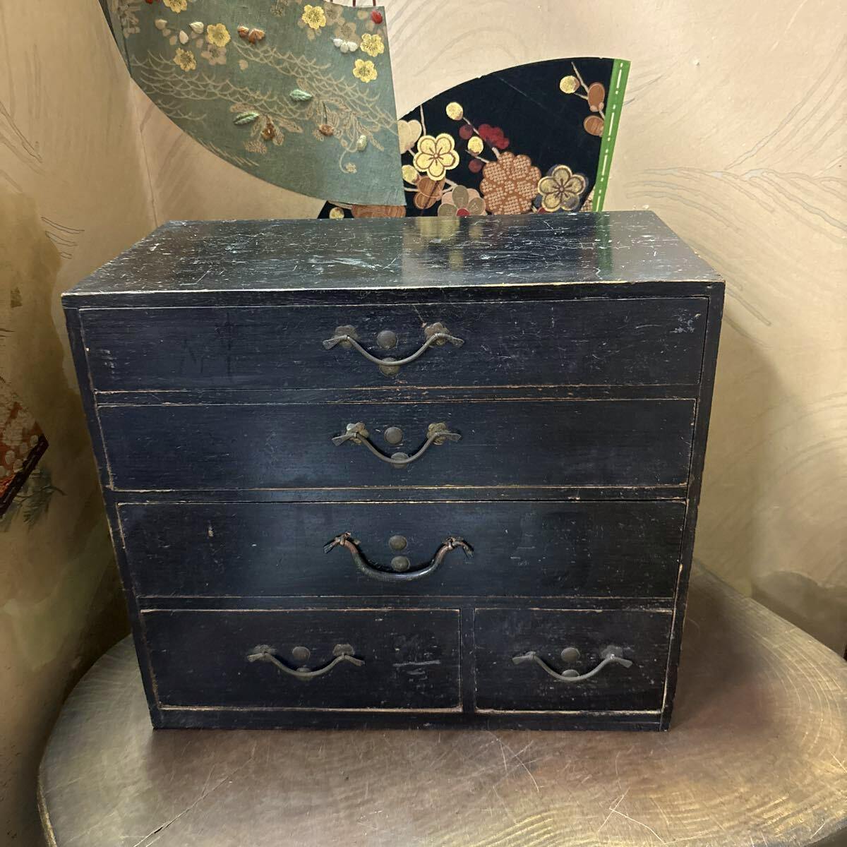 Front view of a small antique wooden chest of drawers with black finish and metal handles placed on a wooden surface.