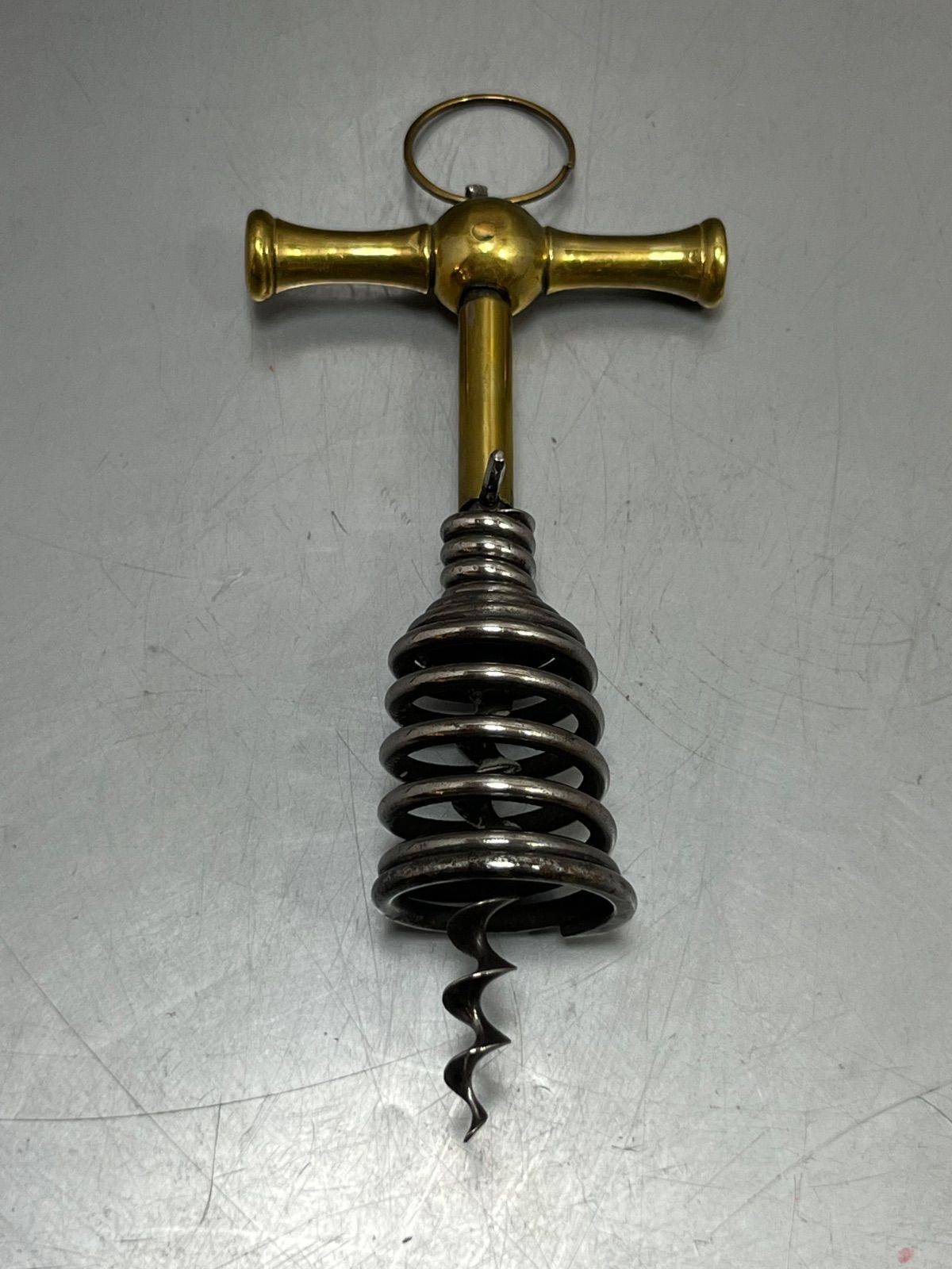 Antique spring-assist wine corkscrew with a brass T-handle and spiral steel spring shown on a gray surface.