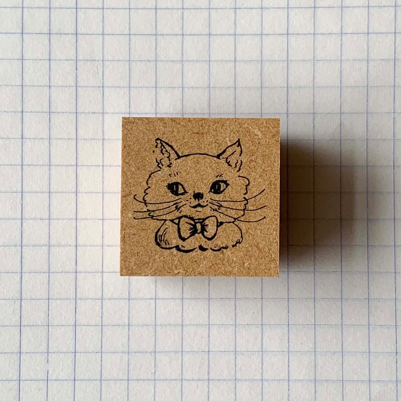 Top view of a square wooden stamp showing a cat face with a ribbon, placed on graph paper.