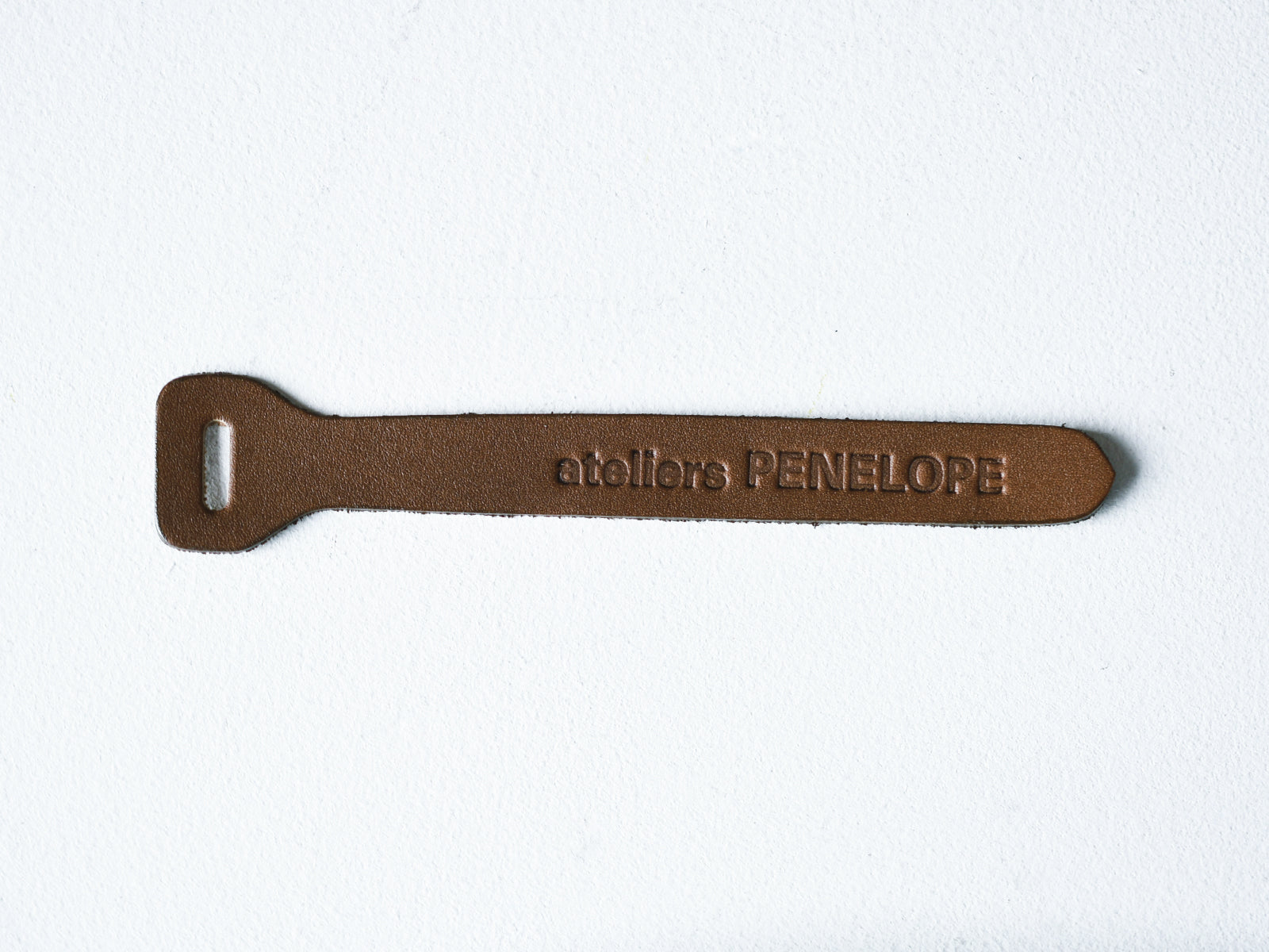 Brown cow leather charm slider embossed with "ateliers PENELOPE" on a white background.