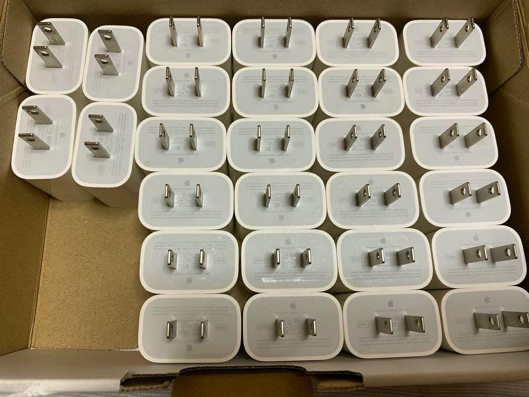 Multiple white Apple USB-C power adapters arranged in a cardboard box, showing their US plug prongs.