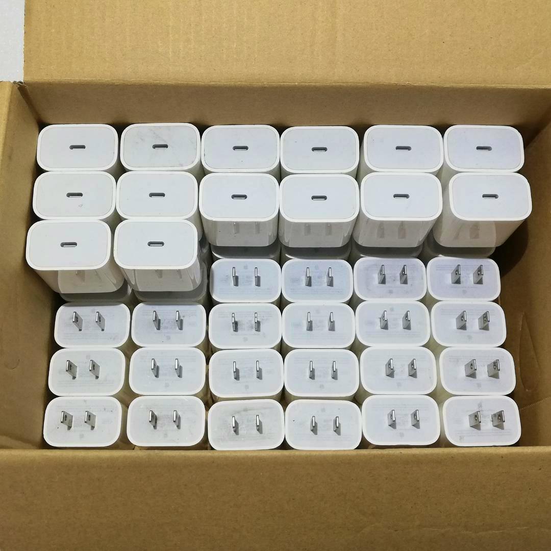 A cardboard box filled with multiple white Apple A2305 20W USB AC adapters arranged in neat rows.