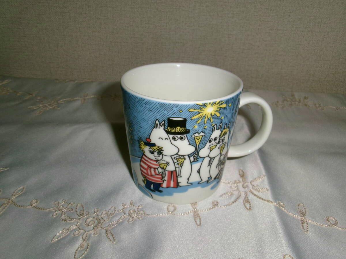 White ceramic ARABIA Moomin mug with characters holding glasses on a blue background, shown angled with the handle on the right and the empty interior visible.