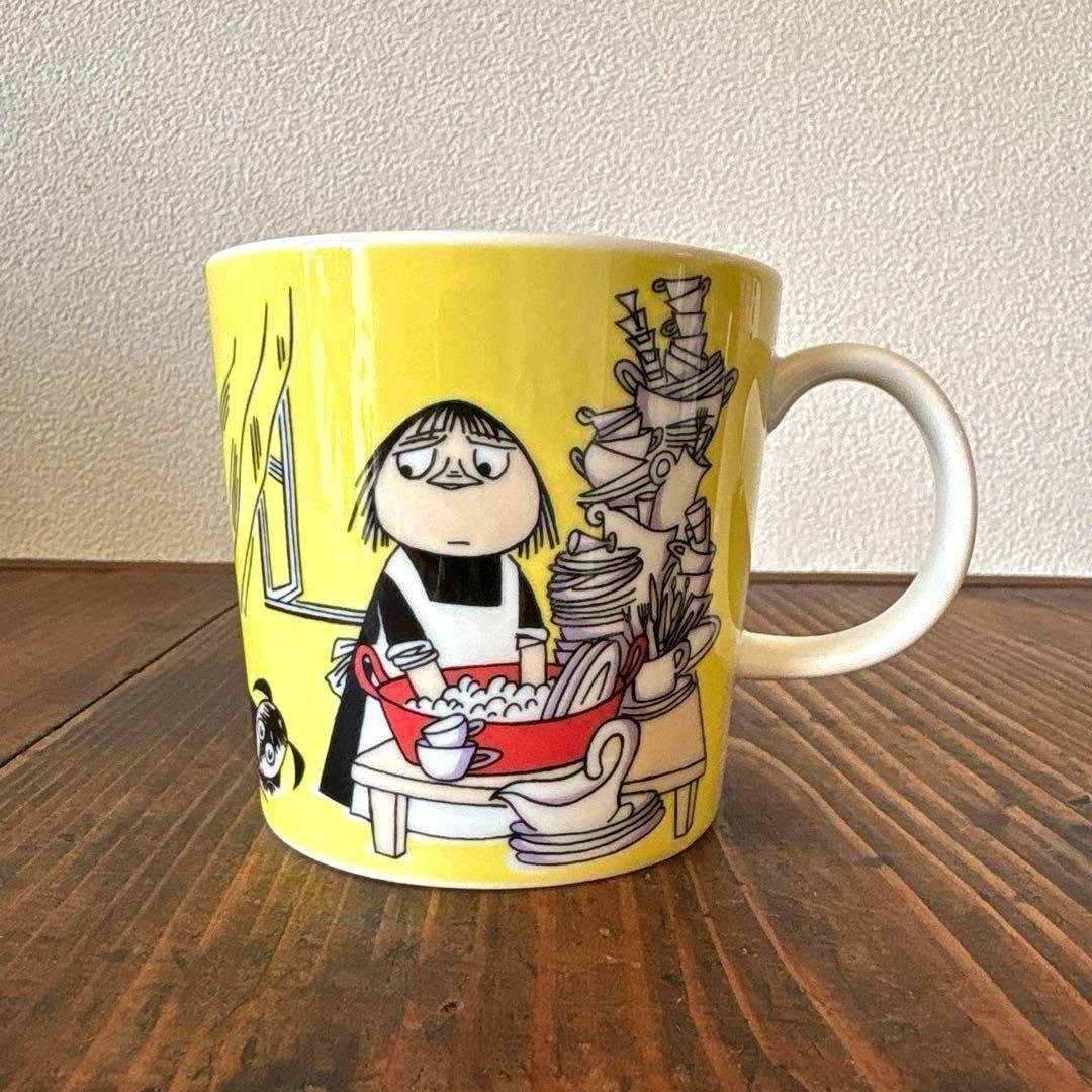 A yellow ARABIA Moomin mug showing the character Misa with dishes, photographed on a wooden surface.