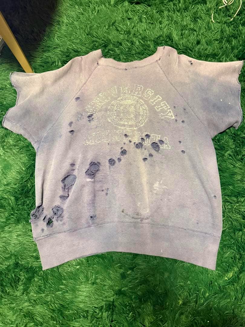Front view of a men's vintage cotton T-shirt with a faded printed logo and multiple distressed holes laid flat on a green rug.