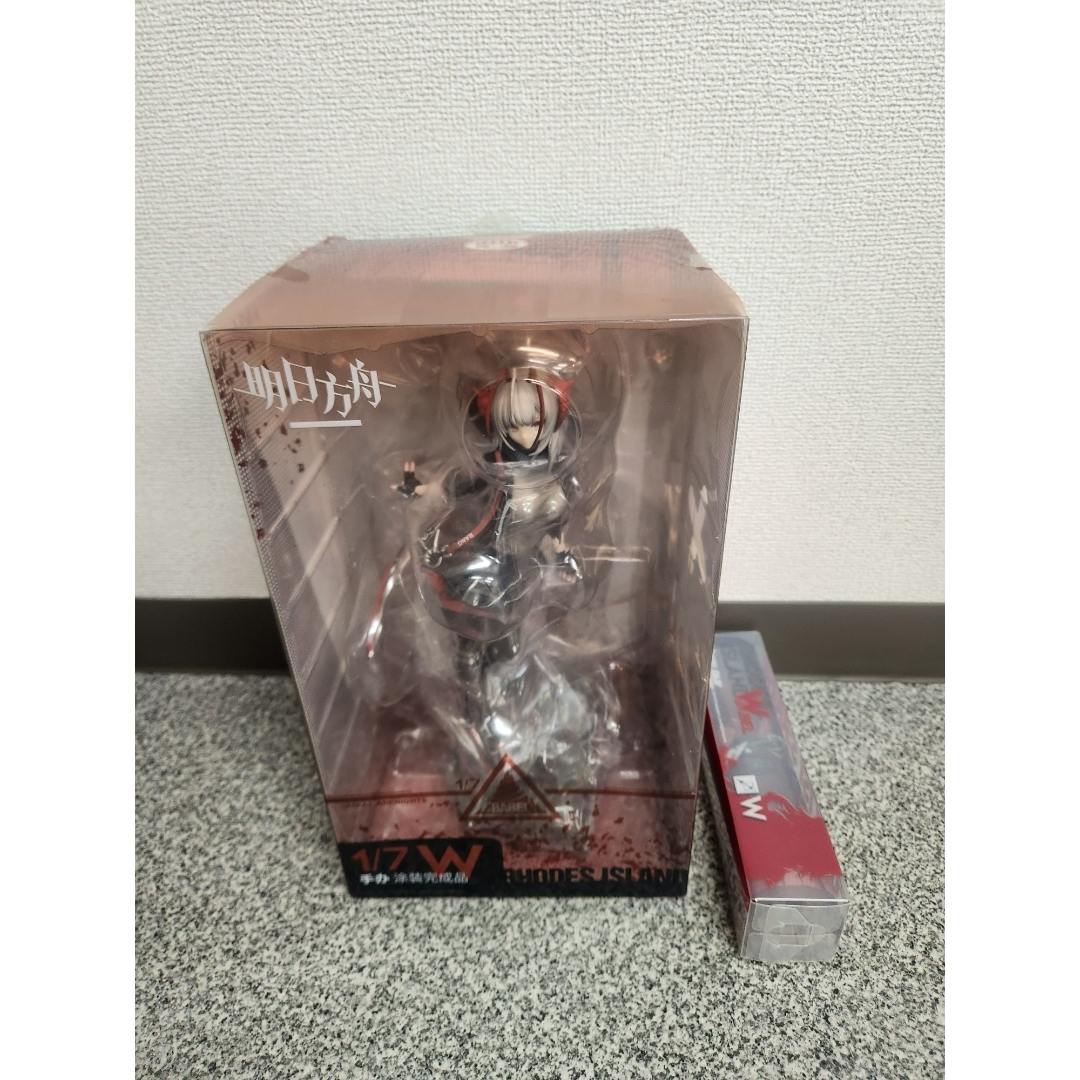 Front view of the boxed Arknights W 1/7 figure visible through the clear window with a small rectangular bonus box beside it on a speckled surface.