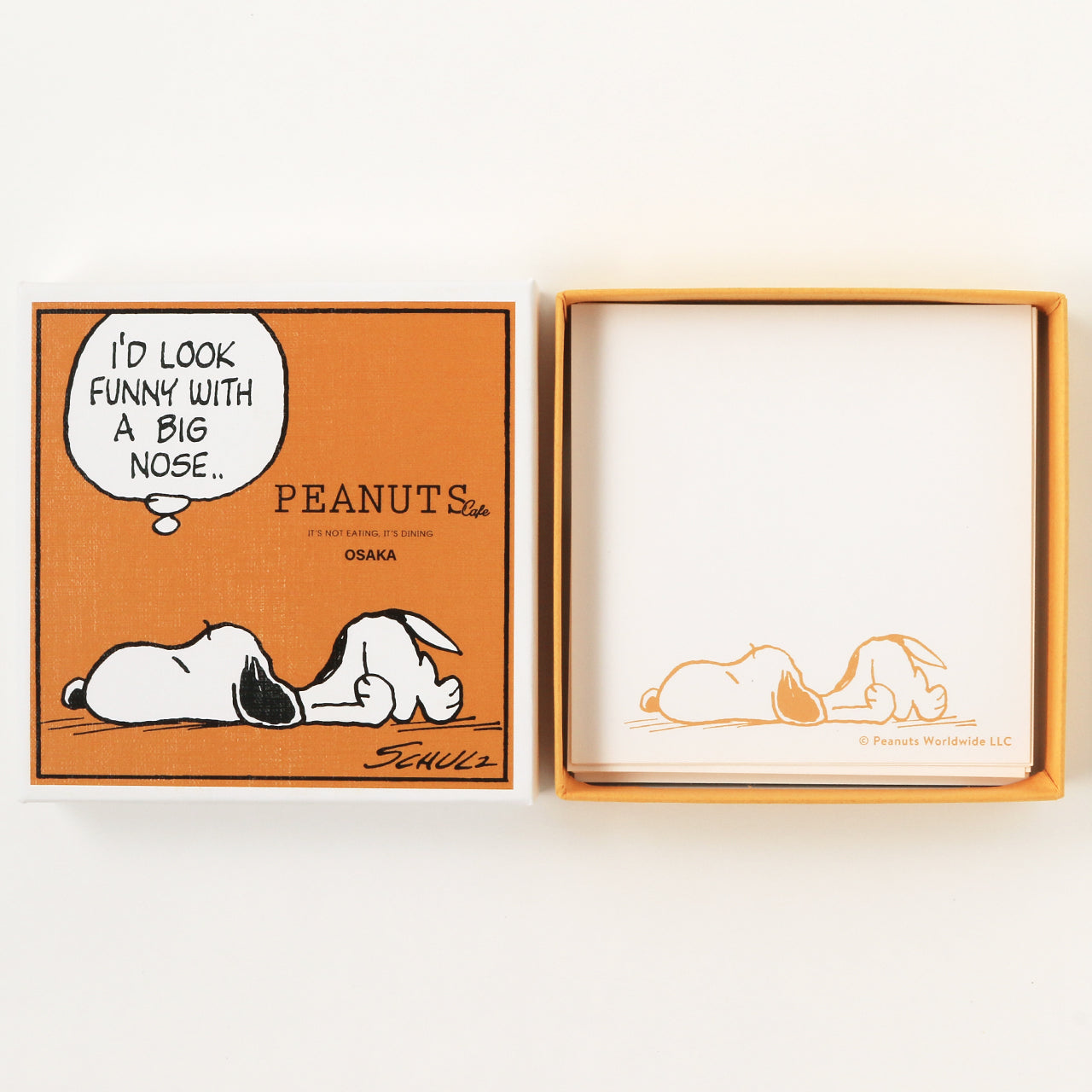 Square art box with a Snoopy illustration on the lid shown next to the open box containing white paper sheets printed with a Snoopy design.