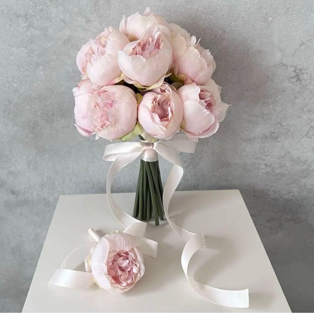 Round artificial wedding bouquet of pale pink roses and peonies with a long ribbon and a matching boutonniere on a white surface.