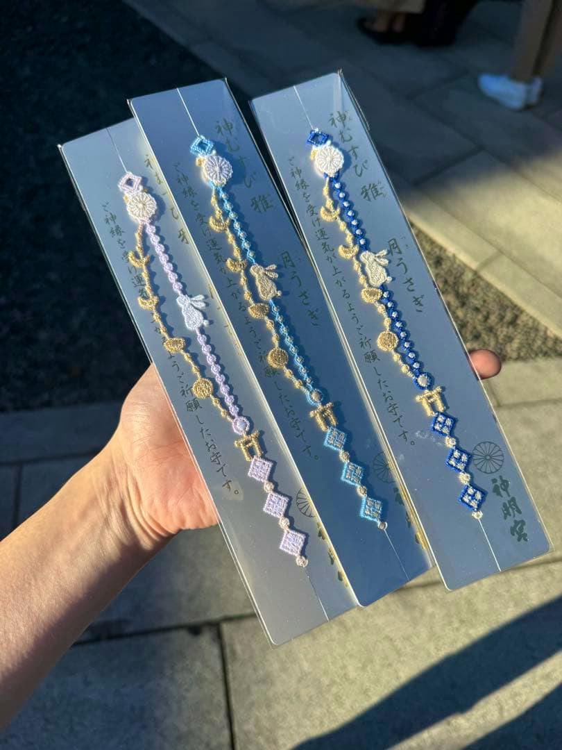 Hand holding three packaged braided amulets on rectangular backing cards, each showing moon and rabbit motifs in light blue, blue, and lavender with gold thread.