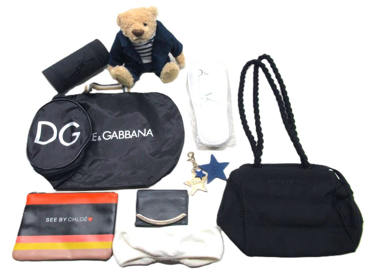 Flat lay of assorted branded small items including a black canvas bag with braided handles, a DG dust bag, a SEE BY CHLOÉ pouch, slippers, and a teddy bear.