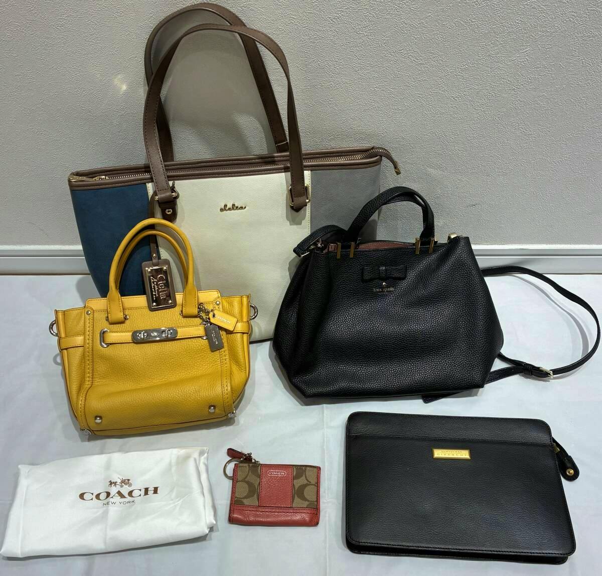 A group of brand bags and accessories arranged on a surface, including a blue-and-white tote, a yellow handbag, a black Kate Spade bag, a Coach dust bag with a small card case, and a black leather pouch.