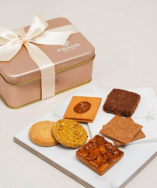 Ribbon-tied metal tin next to six sablé cookies arranged on a marble serving board.