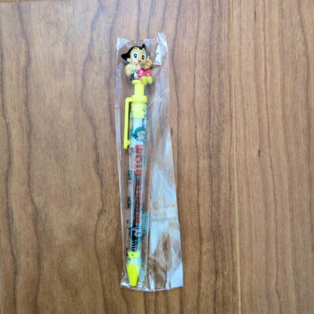 A 鉄腕アトム (Astro Boy) ballpoint pen with yellow accents and a character topper, in a clear plastic sleeve on a wooden surface.