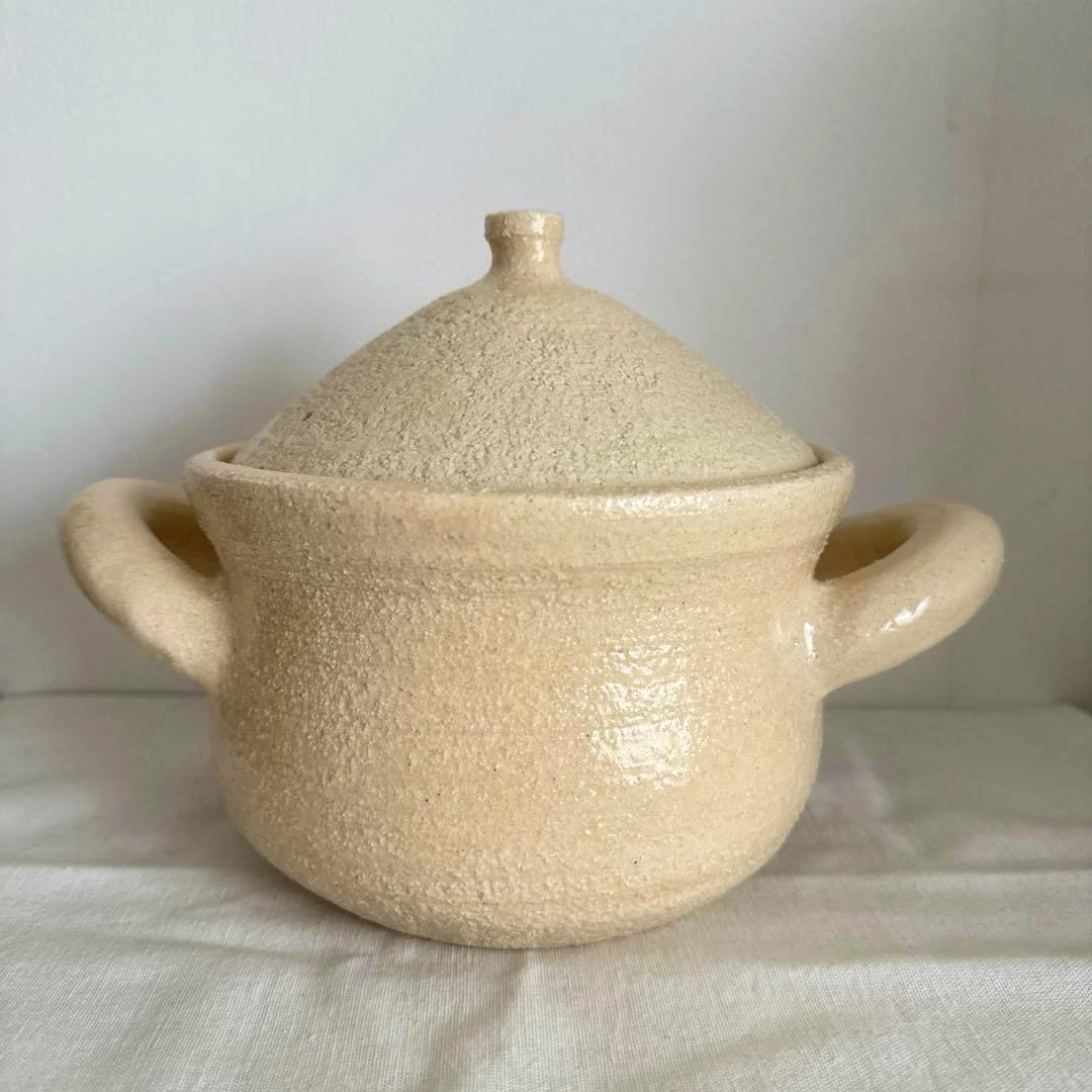 Beige ceramic donabe (clay pot) with lid and two handles on a white cloth surface.