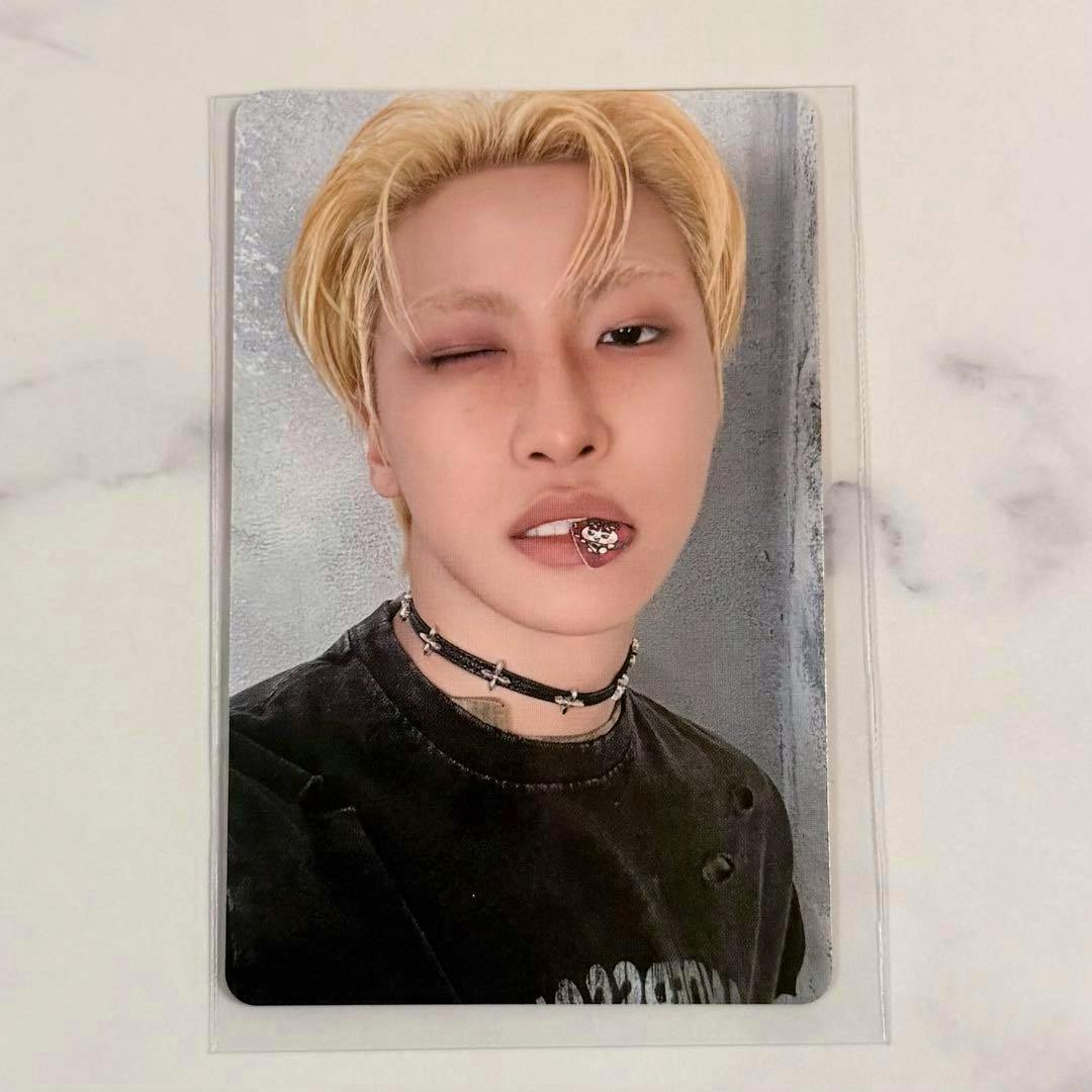 Front of the ATEEZ MIGHTEEZ photocard showing a close-up portrait of the member with blond hair, wearing a choker and a small decoration on the lip, in a clear sleeve on a light surface.