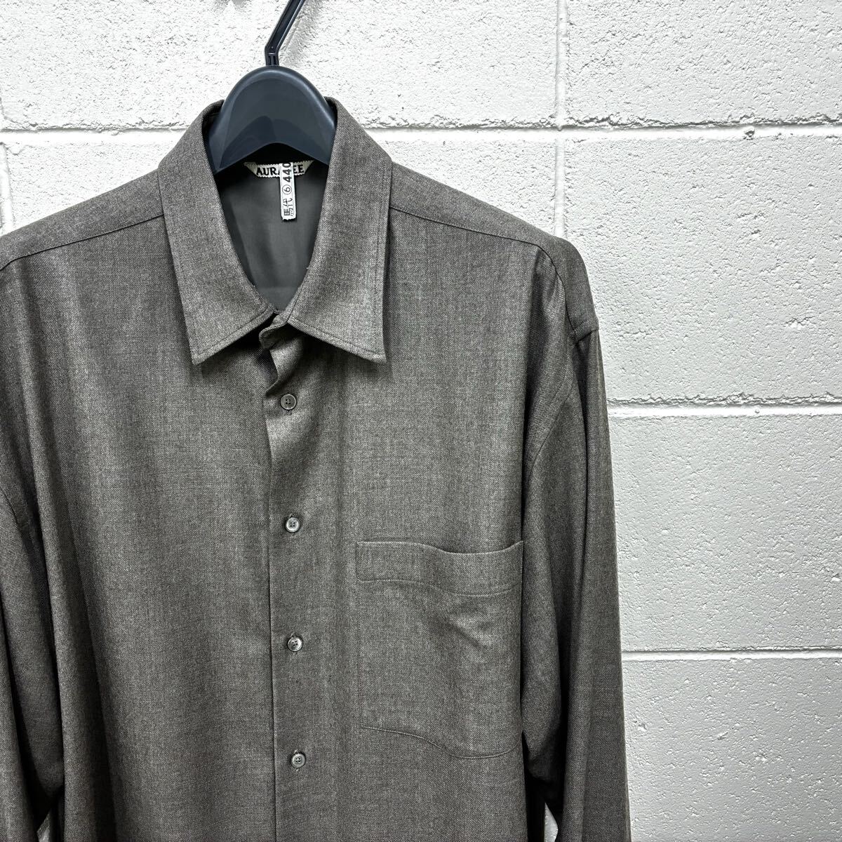 Light brown long-sleeve wool shirt on a hanger against a white textured wall.