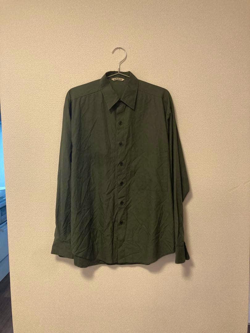 Front view of an AURALEE WASHED FINX TWILL BIG SHIRT in green hanging on a hanger against a plain wall.