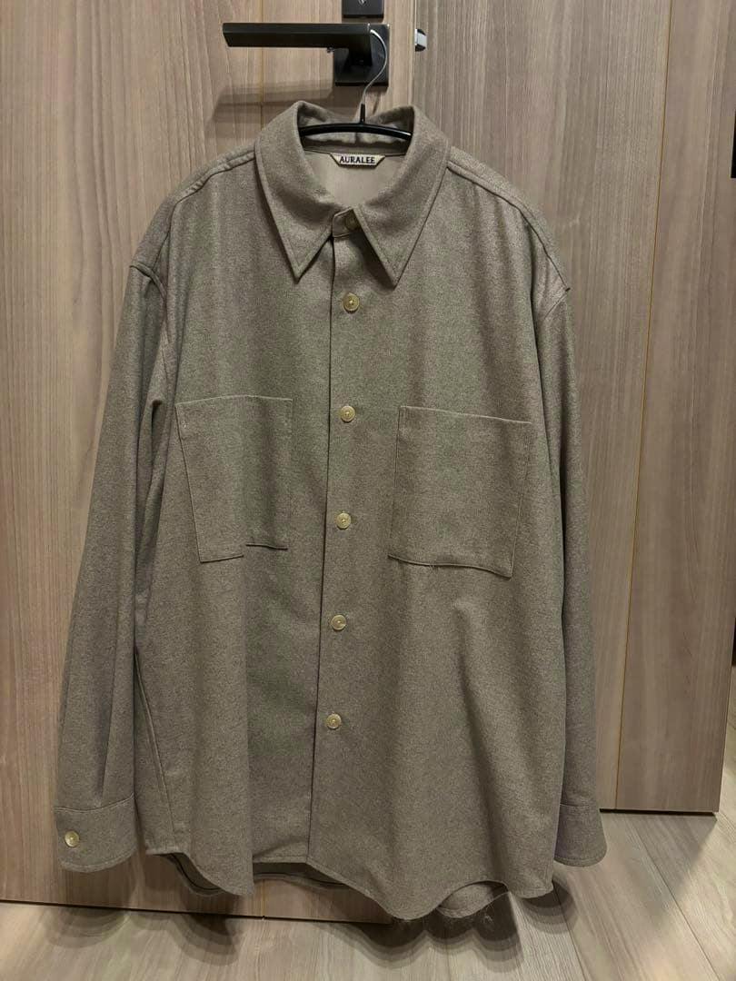 Front view of an Auralee wool fulling flannel shirt hanging on a hanger against a wooden background.