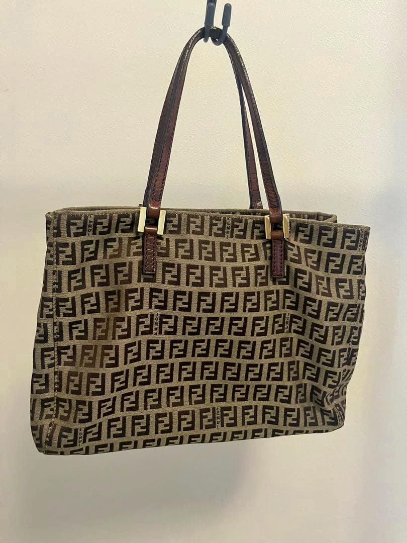 Fendi B5-compatible bag with all-over FF logo print and brown leather handles hanging against a wall.