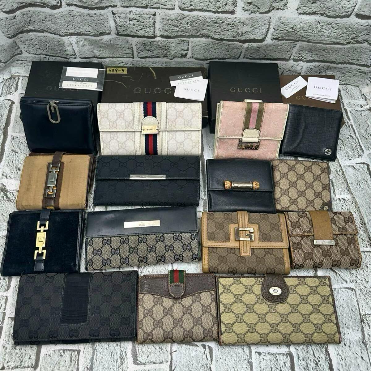 Fifteen Gucci wallets arranged on a textured surface with several Gucci boxes visible, showing various GG canvas and leather long, bi-fold, and tri-fold styles.
