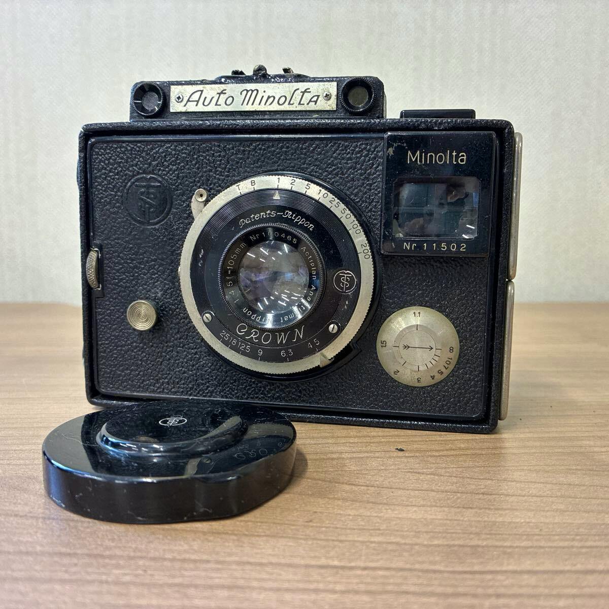 Front view of an Auto Minolta film camera showing the Crown 105mm lens and a detached lens cap on a wooden surface.