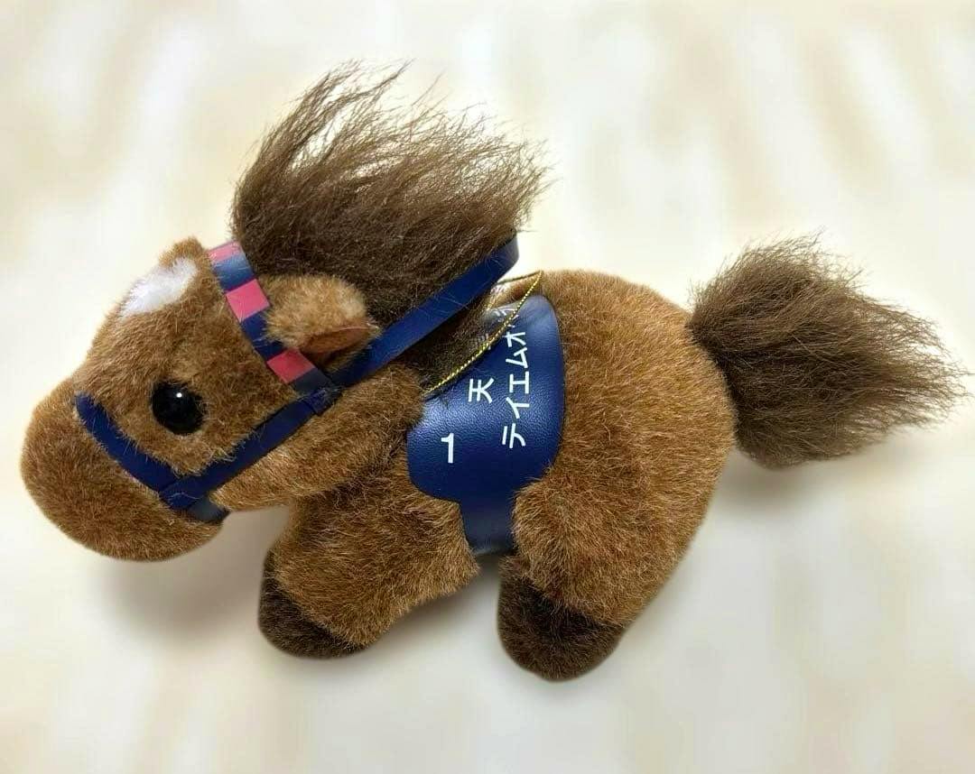 Brown horse plush toy wearing a blue saddle with Japanese writing, photographed on a white background.