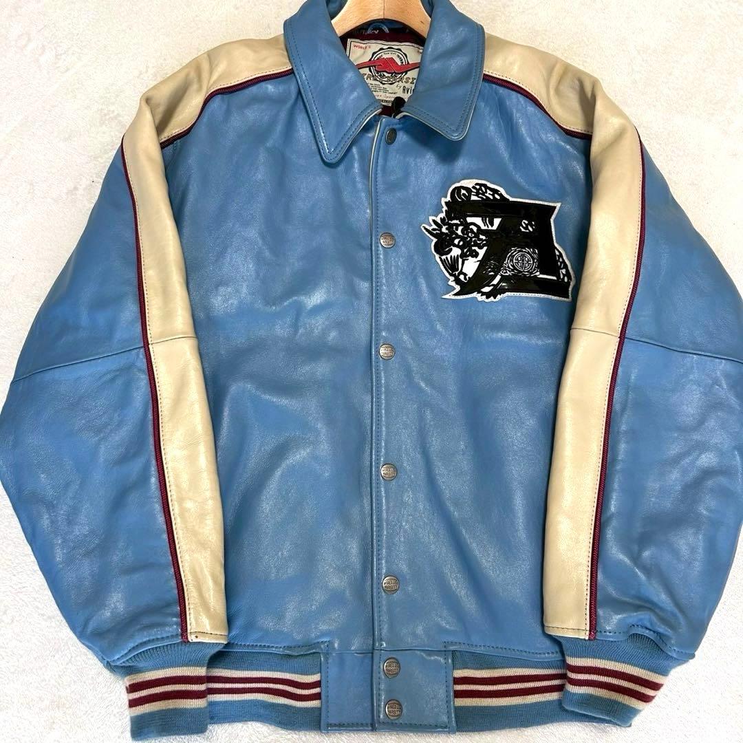 Front view of a men's cow leather Avirex varsity jacket showing a blue body, cream sleeves, snap-front closure, ribbed hem, and a chest embroidered patch.