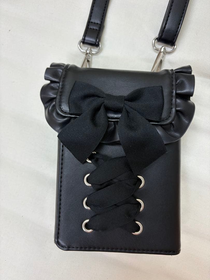 Front view of a black synthetic-leather smartphone wallet shoulder bag with ruffle flap, bow, lace-up eyelets, and shoulder straps attached.