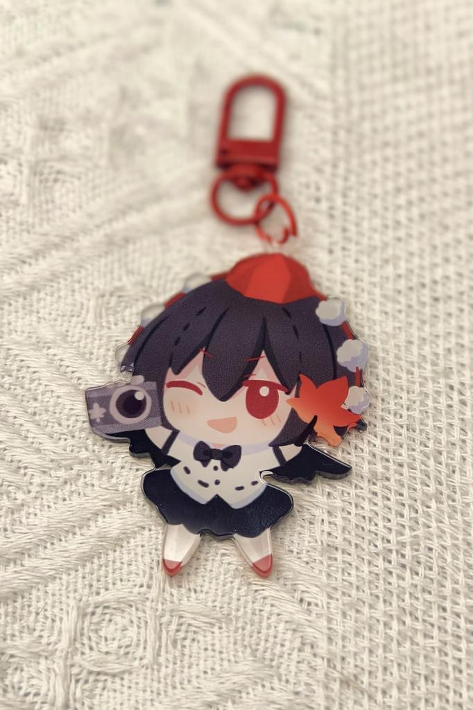 Close-up of a chibi 射命丸文 acrylic keychain with a red clasp, showing the character winking and holding a camera on white knitted fabric.