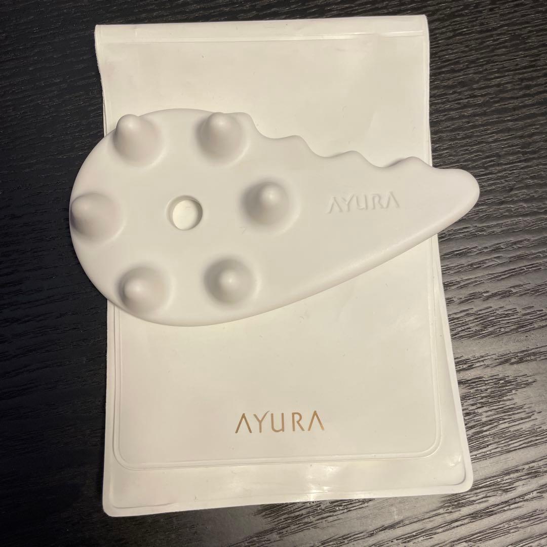 White AYURA facial gua sha massager placed on its white pouch on a dark wood surface.