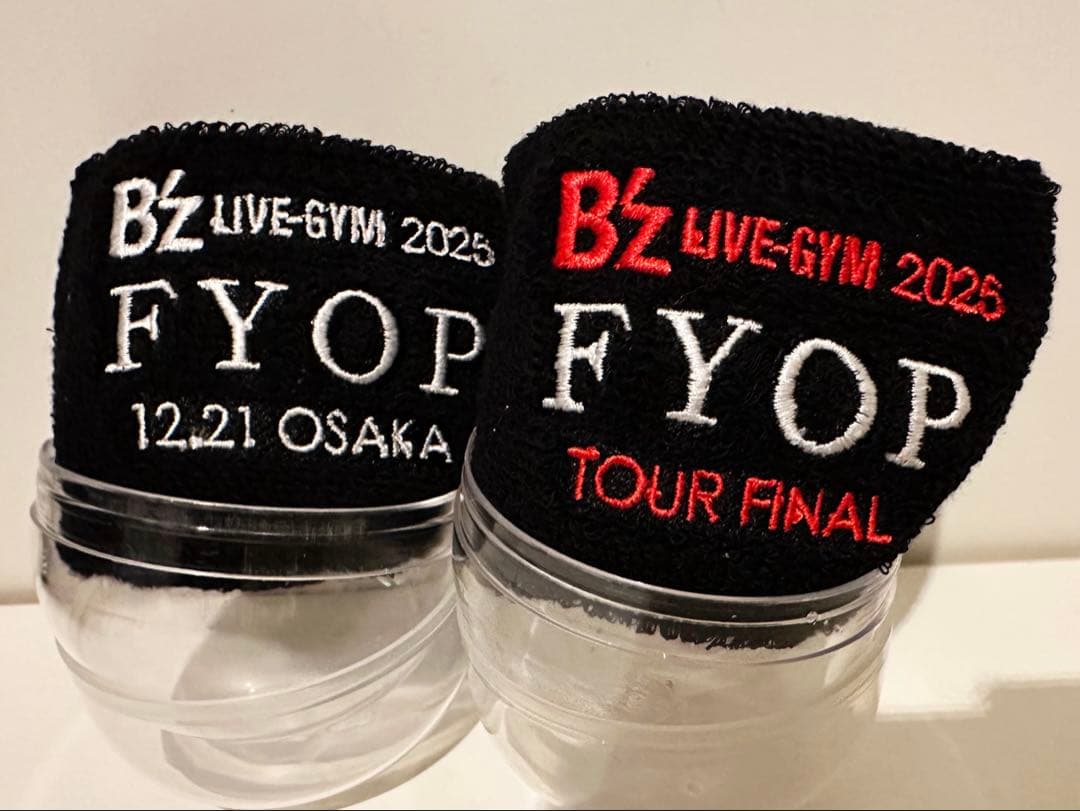 Two black cotton wristbands showing embroidered "B'z LIVE‑GYM 2025 FYOP" and date/tour text, displayed on clear plastic stands.