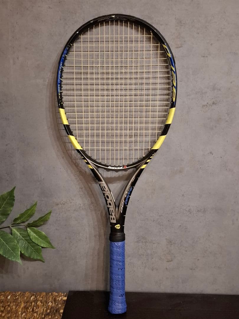 Front view of the Babolat AeroPro Drive G3 (Nadal model) tennis racket with a blue grip leaning against a gray wall.