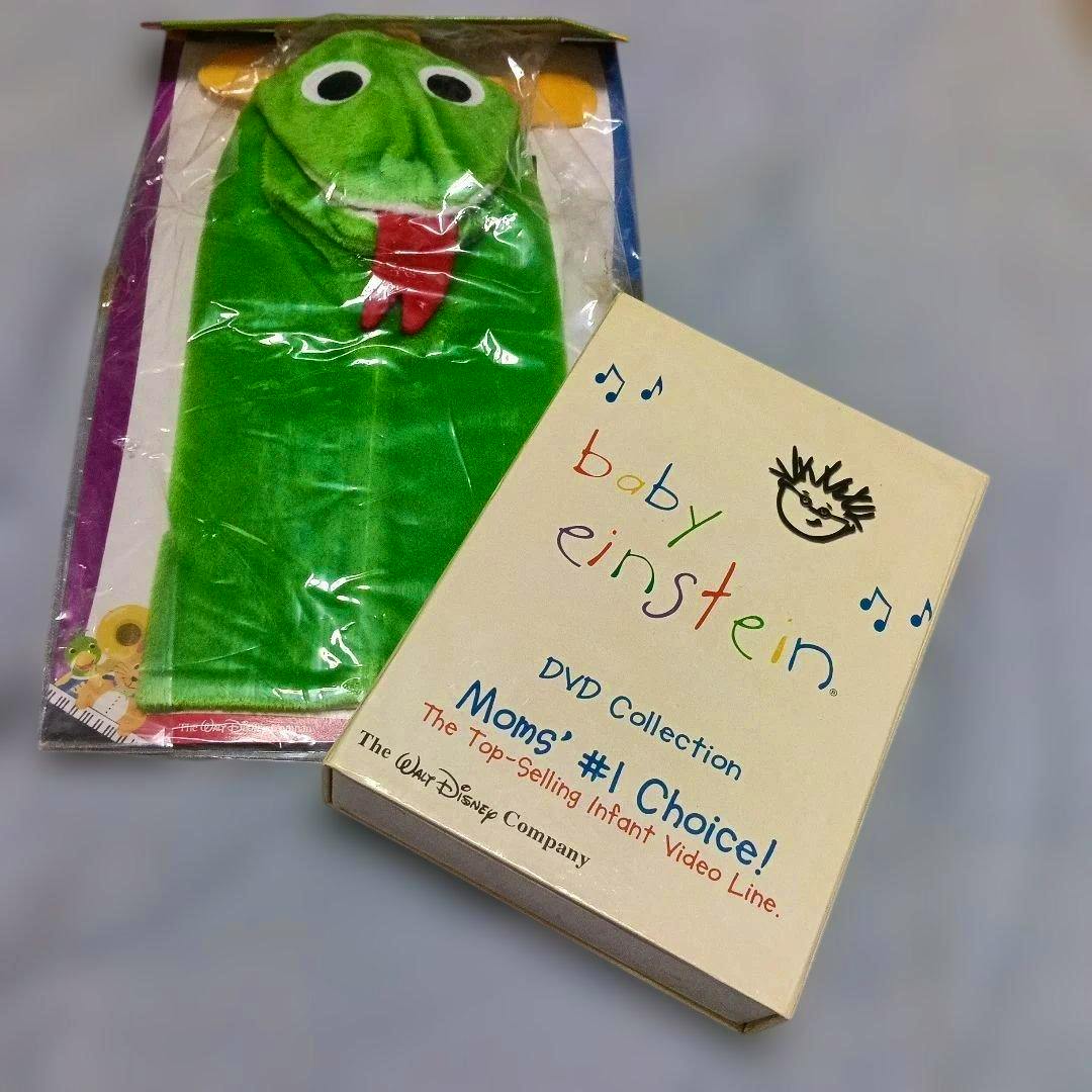 A Baby Einstein DVD collection box pictured next to a green plush puppet in plastic packaging.