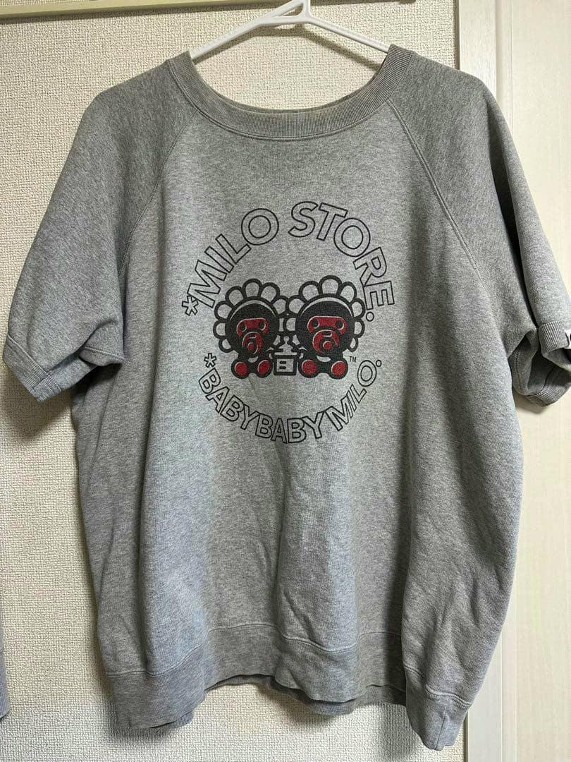 Gray short-sleeve sweatshirt (cotton) hanging on a hanger showing a centered BABY MILO STORE graphic on the front.
