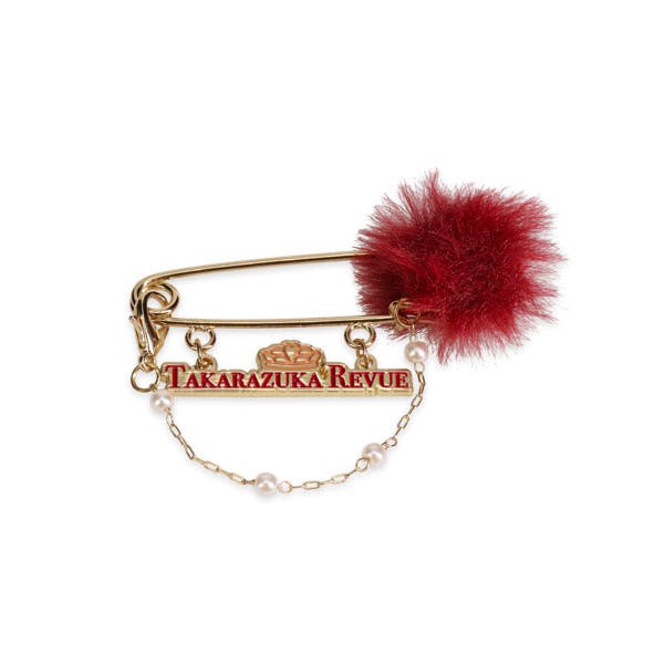 Gold-tone metal bag/stole pin brooch with a red fur pompom, a dangling "TAKARAZUKA REVUE" bar, and a small pearl chain.