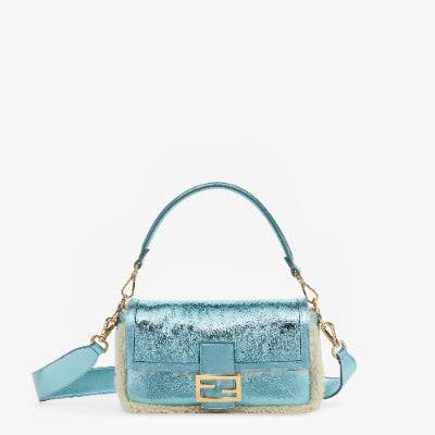 Front view of a metallic teal lambskin leather baguette bag with a top handle, detachable shoulder strap, shearling trim, and gold-tone buckle on a white background.