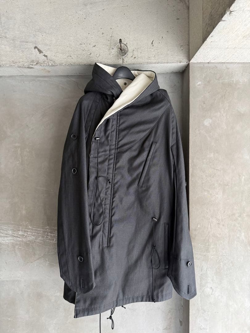 A men's bajra Archive multi-drawstring cotton woven coat hanging on a hanger against a concrete wall.