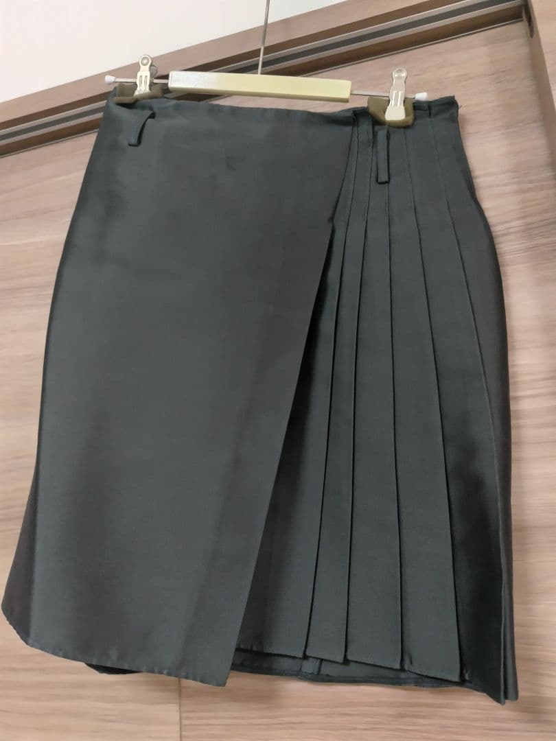 Front view of a black knee-length silk Balenciaga pleated skirt hanging on a hanger, showing a wrap panel and knife pleats.