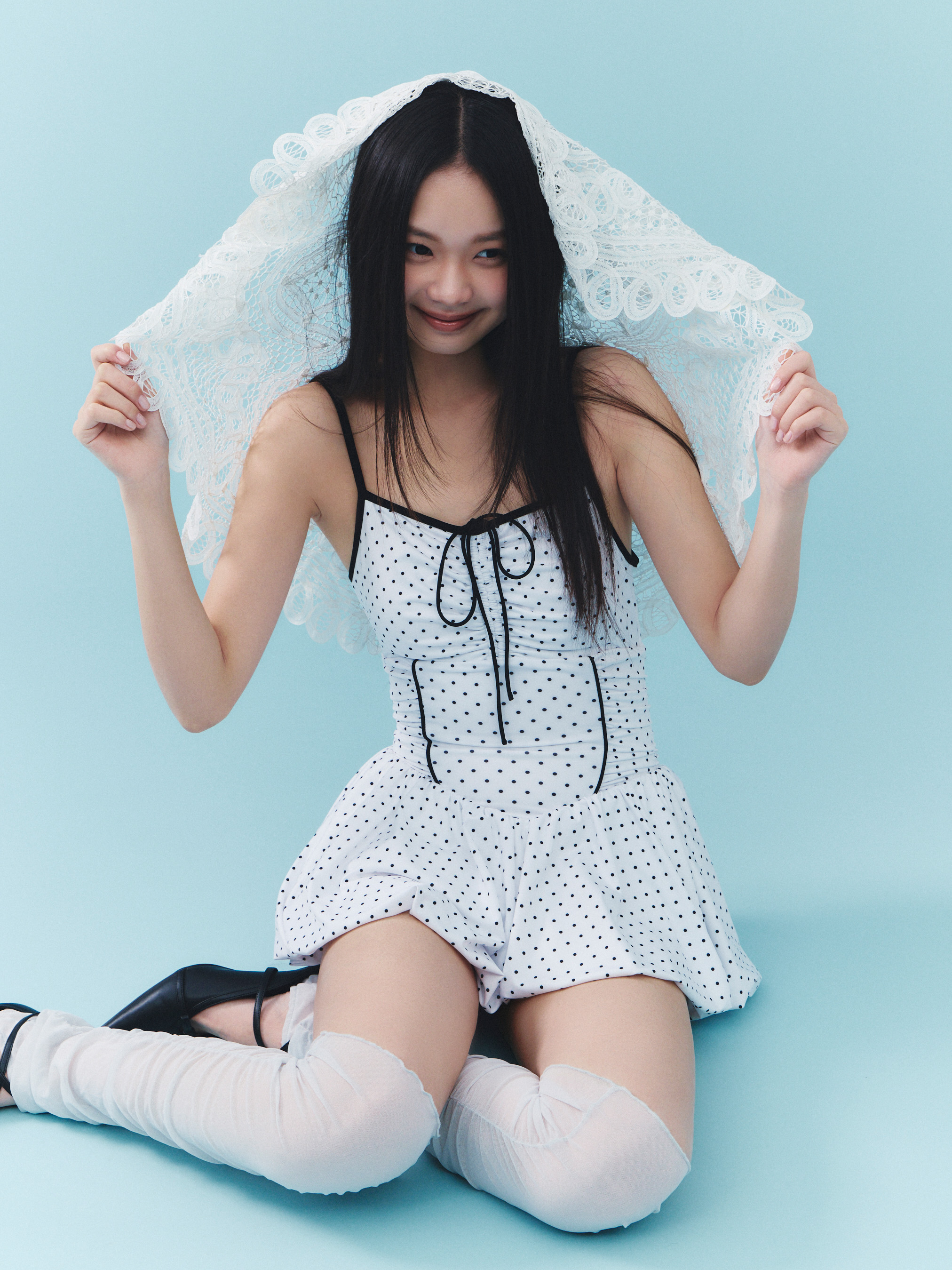 Model wearing a white polka-dot balloon dress-style swimsuit kneeling against a light blue background while holding a lace shawl over her head.
