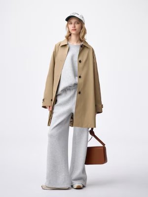 Female model wearing a beige A-line half coat over a gray outfit, holding a brown handbag and wearing a cap.
