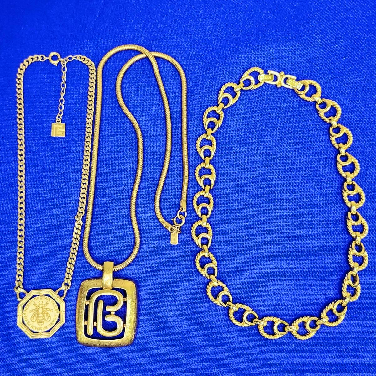 Three gold-colored Balmain necklaces arranged on a blue fabric background, showing pendants and chain links.