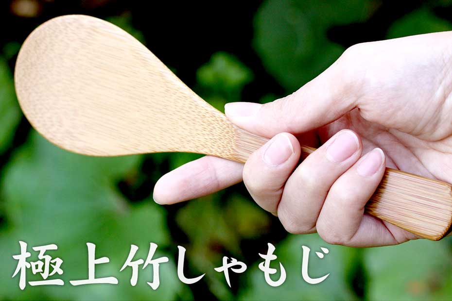 A bamboo rice paddle (wooden spoon) held in a hand against a blurred green background.