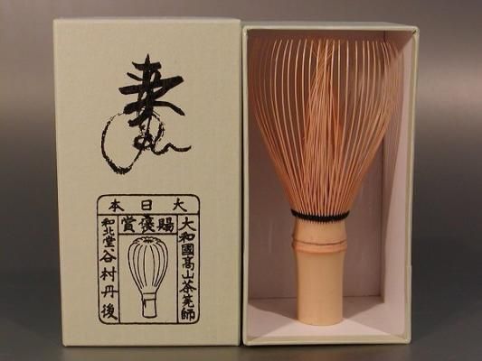 A bamboo tea whisk (chasen) standing upright inside a rectangular box next to a box lid printed with Japanese characters.