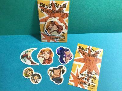 Photograph of illustrated stickers arranged on a blue surface alongside the 'BANG! BANG! STICKERS' product packaging and an informational card.